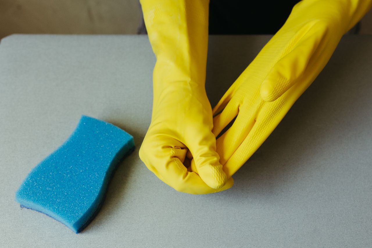 services-01 Vue rapprochée de mains portant des gants jaunes à côté d’une éponge bleue sur une surface grise.